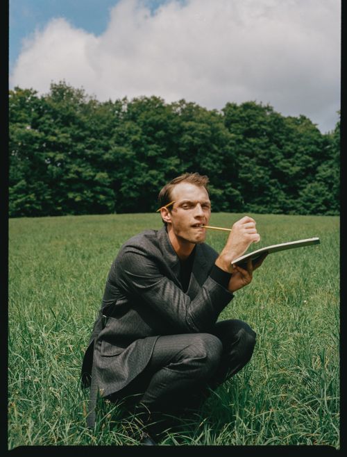Man in suit squats in grassy field, holding pencil to notepad, forest background, blue sky and clouds above, outdoor setting.