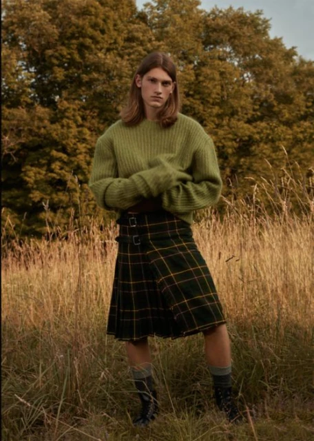 Man in kilt standing in grassy field with trees. He wears a green sweater, kilt, and boots. Outdoor fashion shot, medium shot.