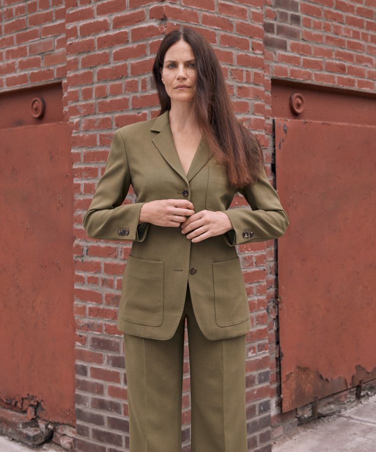 Adult woman with long brown hair poses in front of a brick wall wearing a green, two-button pantsuit with wide-leg pants and patch pockets.