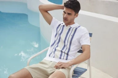A man wearing a white polo shirt with blue stripes and beige shorts seated on a poolside chair in Los Angeles.
