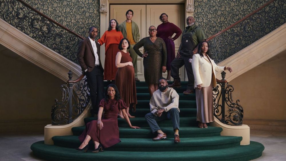 Group of diverse adults poses on a staircase indoors. The individuals wear various earthy-toned outfits in a portrait-style photograph.
