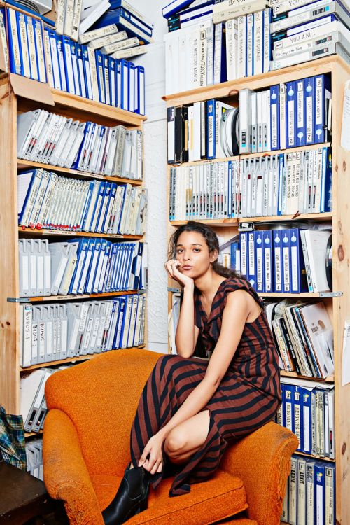 A young woman in a striped dress and black boots sits in an orange chair in a room with floor-to-ceiling bookshelves filled with binders.