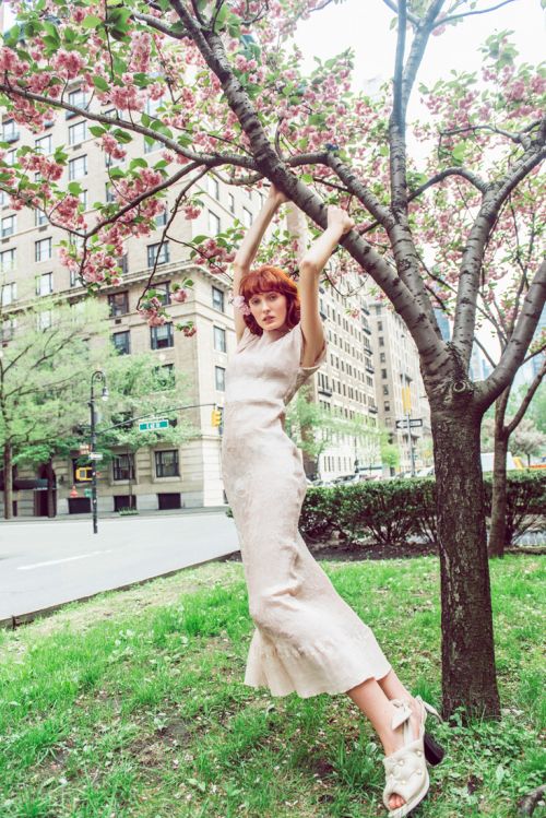 A slender woman with auburn hair in a beige dress hangs from a blooming tree in an urban park setting with buildings in the background.