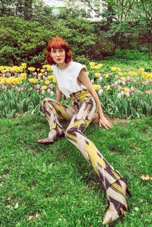 A woman with red hair sits in a flower garden, wearing a white top and patterned pants and boots. The garden has yellow and pink tulips.