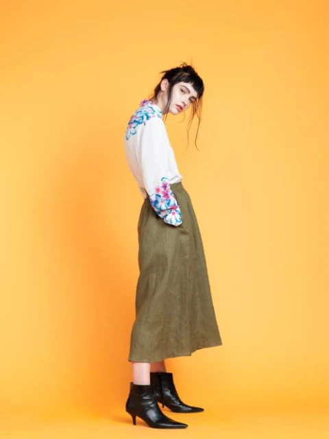 Full-length shot of a young adult woman in an off-white, floral-print blouse and olive green midi skirt, posing against a yellow backdrop.