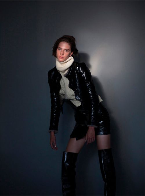 A stylish young woman strikes a pose indoors wearing a black patent jacket, high waisted shorts, and knee-high boots against a dark wall.