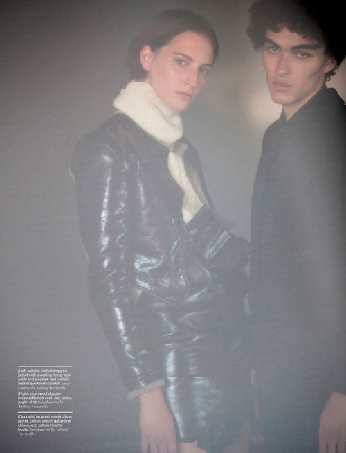 Editorial fashion shot featuring two young adult models wearing dark leather and wool outfits against a soft grey background in studio setting.