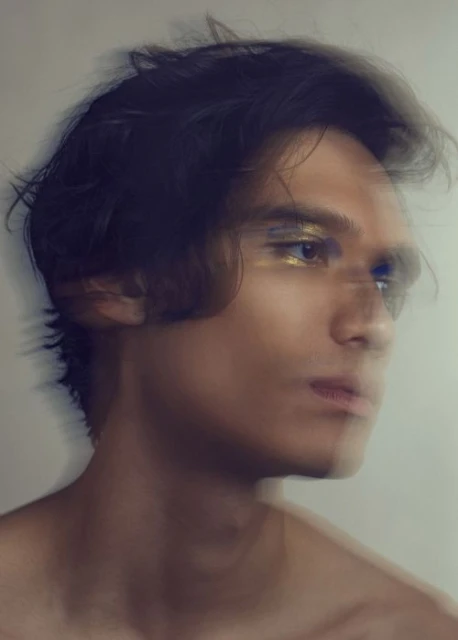 Portrait of a young adult male with dark hair and gold makeup around the eyes, shown in a blurred motion effect against a neutral background.