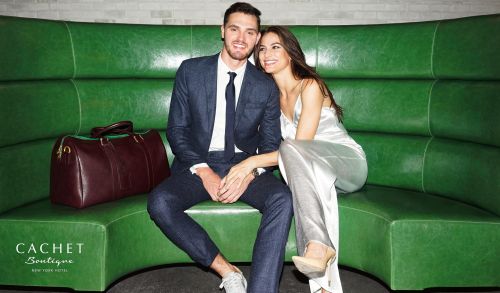 Well-dressed couple poses on green leather banquette with a burgundy duffel bag, promoting a hotel boutique.