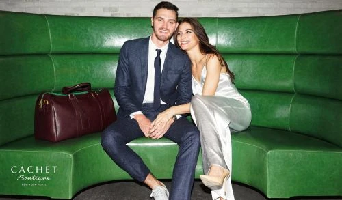Well-dressed couple poses on green leather banquette with a burgundy duffel bag, promoting a hotel boutique.
