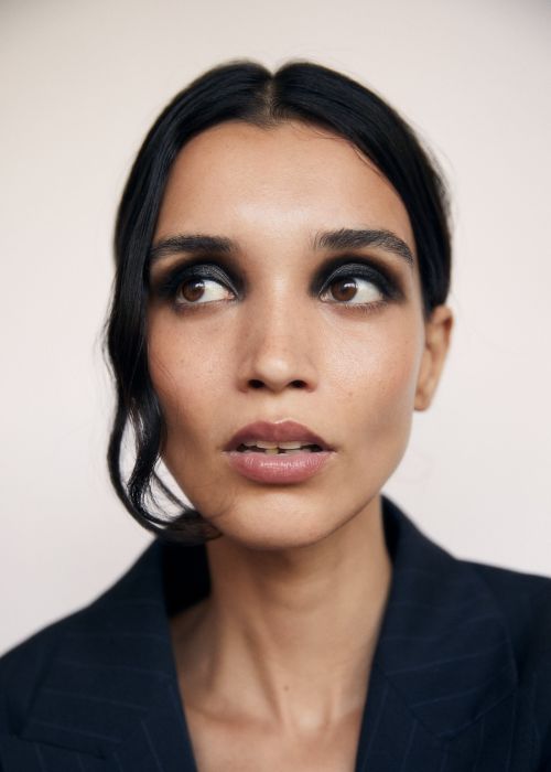 Close-up portrait of a woman with dramatic black eye makeup wearing a dark pinstripe suit jacket against a light background.