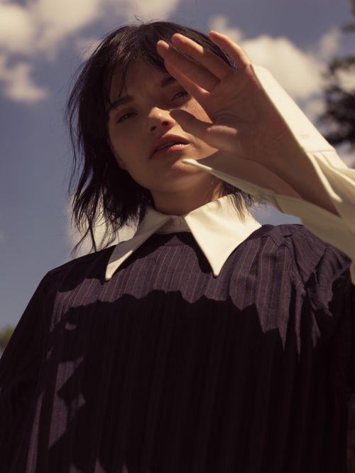Woman with short dark hair shields eyes from the sun in pinstripe jacket with large white collar outdoors with sky and trees.