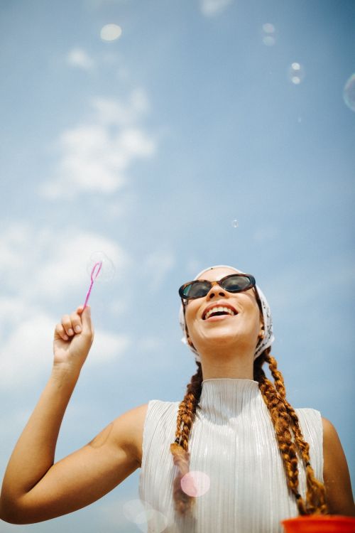 Smiling woman blowing bubbles under a bright blue sky. She has sunglasses, a headscarf, and wears a sleeveless white top.