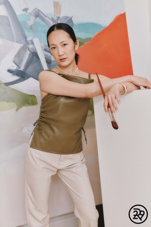 Adult woman in a sleeveless leather top and slacks poses indoors against a painting with a brush in her hand and white background.