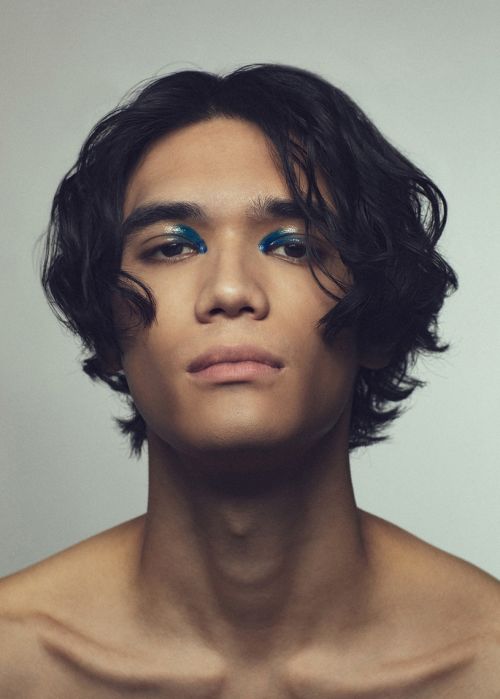 Close-up portrait of a young adult male with dark wavy hair and bold blue eyeshadow on a neutral background in a beauty context.