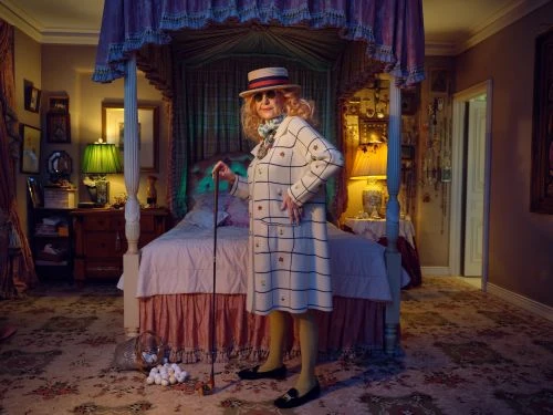 An elderly woman in a patterned coat and hat stands in an ornate bedroom holding a golf club, surrounded by golf balls.