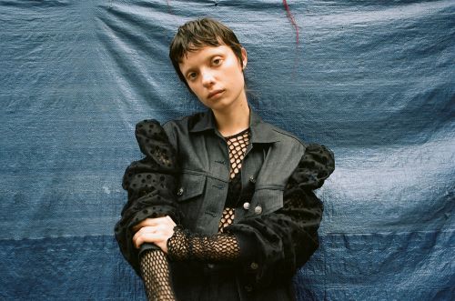 Young adult female posing in a dark denim jacket and fishnet top against a blue textured backdrop in a medium, eye-level studio shot.