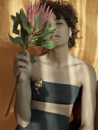 Model with curly hair holding a pink protea flower in front of her face, wearing a dark bandeau top and gold brooch.