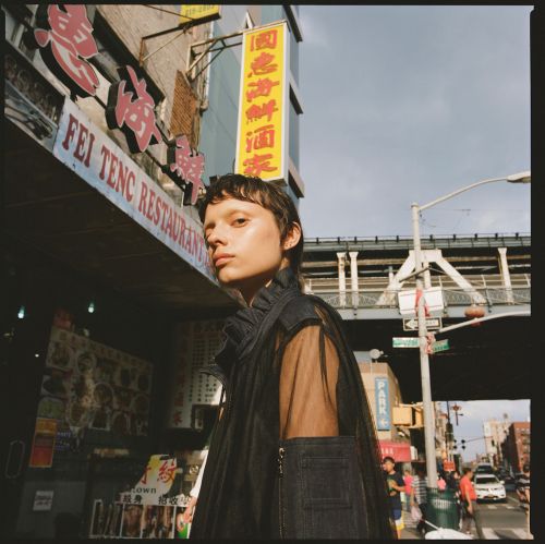 Adult female model in sheer, denim-accented top poses in Chinatown with building signs, cloudy skies, street and elevated train tracks.