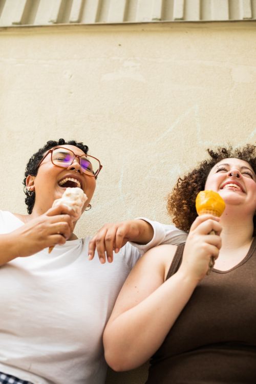 Two smiling women with curly hair are standing side by side outdoors eating ice cream cones with a neutral-colored wall background.