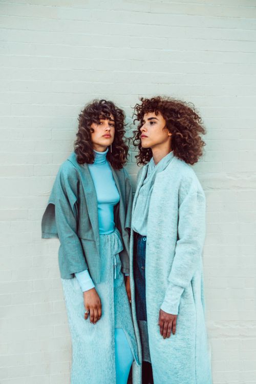 Two women in matching turquoise and blue-grey outfits, long curly hair, in a fashion shot in front of a textured wall. 