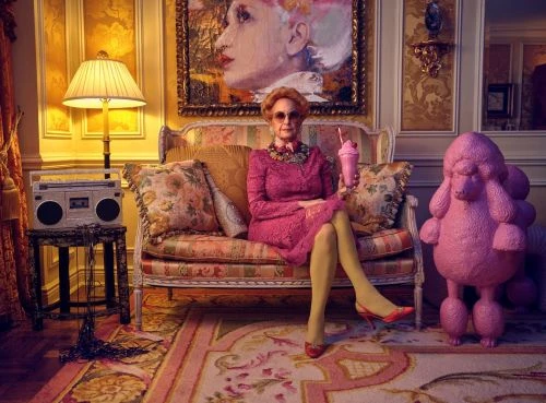 Elegant senior woman in a pink dress sits on an ornate couch, holding a milkshake, with a vintage radio and pink poodle statue.