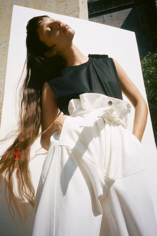 Brunette woman in black sleeveless top and white wide-leg pants poses outdoors with sunlight on her face and body.