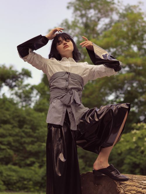 Woman in avant-garde fashion poses on a log in a wooded area. She wears a button-down shirt, black wide-leg pants and black loafers.