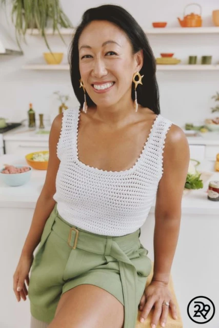 Smiling Asian woman in white top and green shorts poses in a kitchen setting with white cabinets and various cooking items in the background.