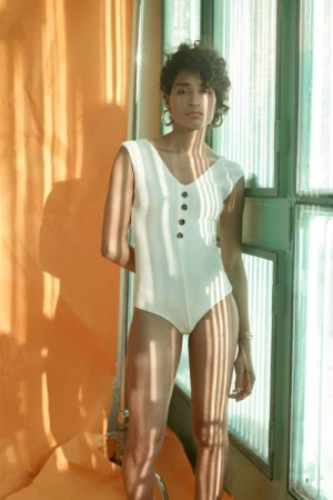 An adult female model poses indoors wearing a white bodysuit with buttons, standing against a window with light filtering through.