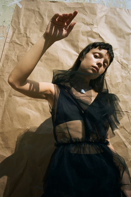 Young adult woman in a black, sheer dress poses with an outstretched arm in front of a tan paper background in a portrait shot.