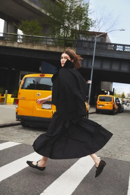 Woman in stylish black dress crossing street with yellow taxi cabs in urban setting, showcasing fashion and city life.