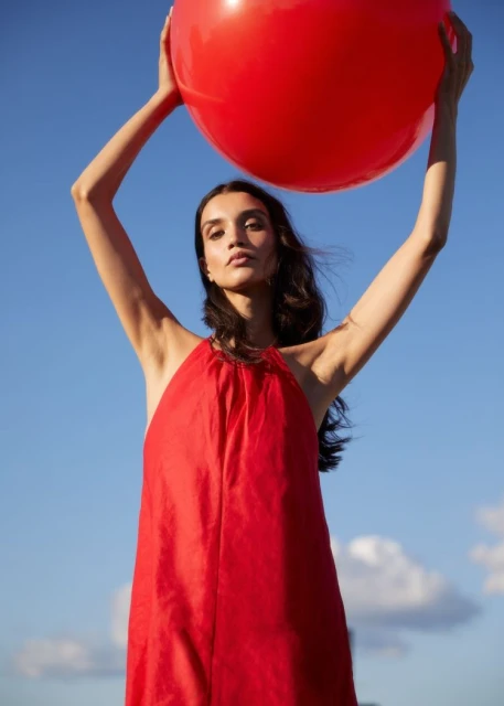 Young woman in red halter dress poses outdoors holding a large red ball against a blue sky; portrait; beauty and fashion concept.