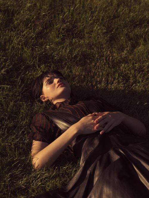 Woman in a black dress laying in a green grass field with soft lighting and shallow depth of field looking up at the sky.