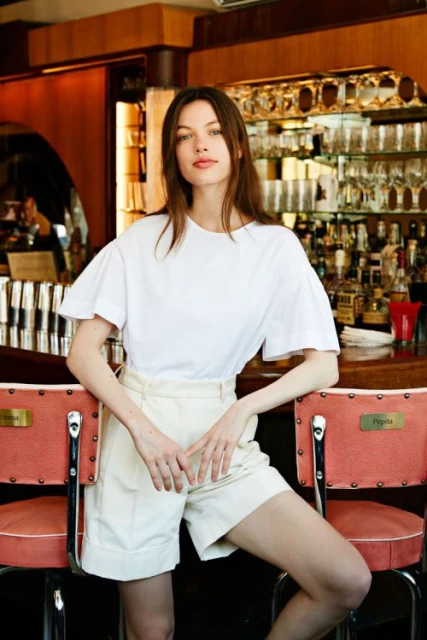 Portrait of a young adult woman with long brown hair posing indoors. She wears a white t-shirt and beige shorts, leaning against red bar stools.