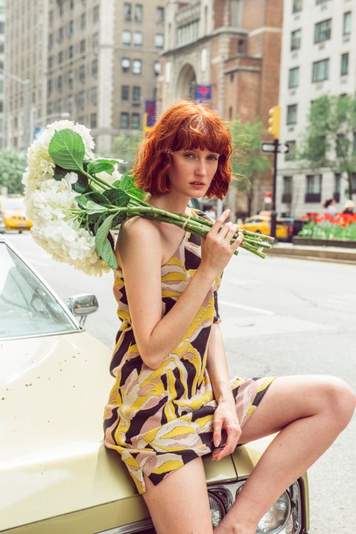 Adult woman with red hair sits on vintage car hood in city street holding flowers. Buildings and yellow cabs are in background.