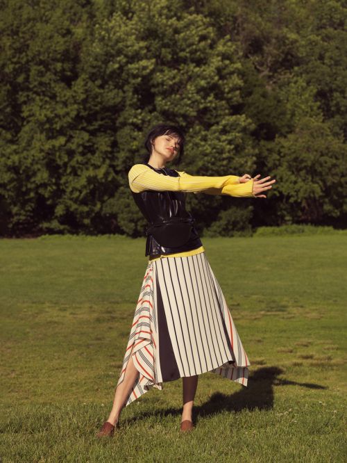 A woman in a yellow sweater and black vest stands on a grassy field in a striped skirt with trees in the background.