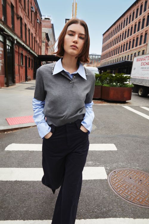 A woman in a gray sweater and black pants walks on a crosswalk in a city, surrounded by brick buildings and urban infrastructure.