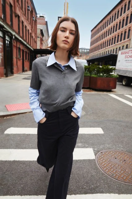 A woman in a gray sweater and black pants walks on a crosswalk in a city, surrounded by brick buildings and urban infrastructure.