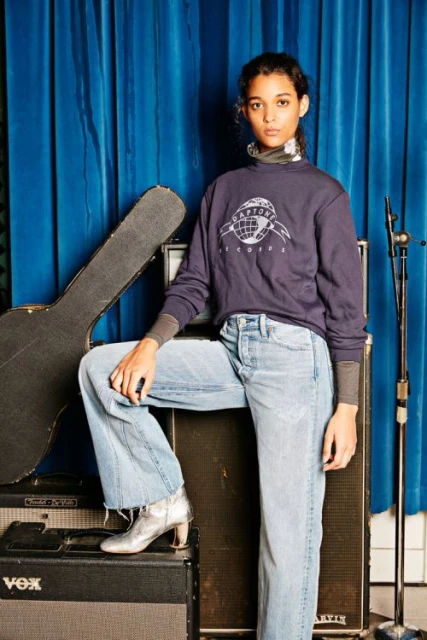 Fashionable woman in a sweatshirt and light-wash denim poses against a blue curtain backdrop with music equipment visible in the background.