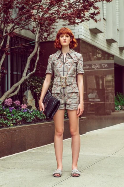 Red-haired woman in patterned romper holding black bag poses on sidewalk, framed by building and tree with red leaves. Fashion shot.