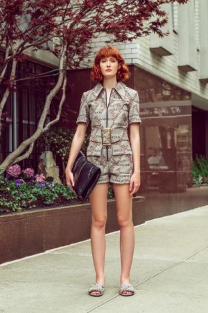 Red-haired woman in patterned romper holding black bag poses on sidewalk, framed by building and tree with red leaves. Fashion shot.