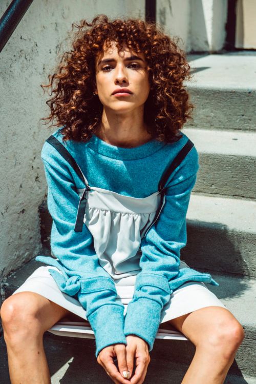 A woman with curly hair sits on concrete steps wearing a blue sweatshirt and white shorts, staring at the camera in natural daylight.