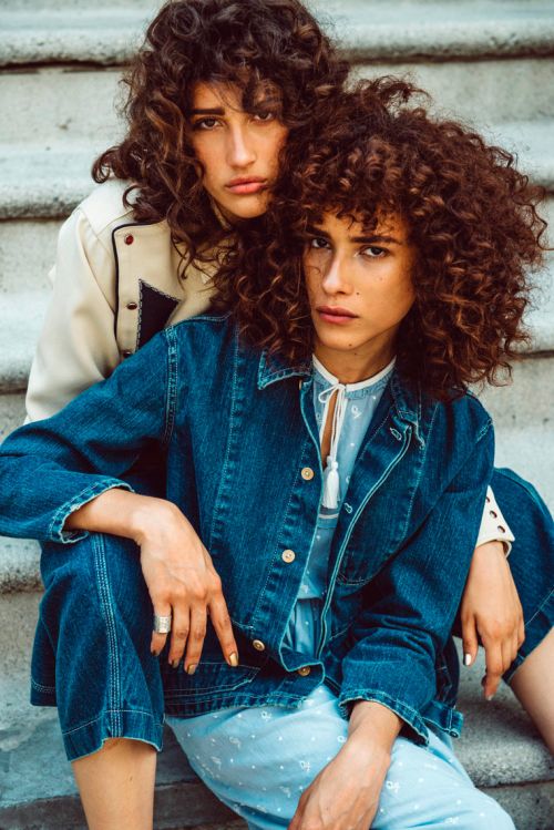 Two women with curly hair pose on stone steps in denim and light-colored jackets, showcasing an outdoor fashion editorial look.