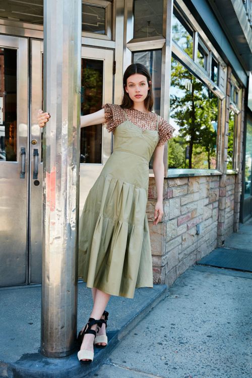 A young woman in a green dress poses in front of a building with metal doorframes. She is standing next to a silver pole.
