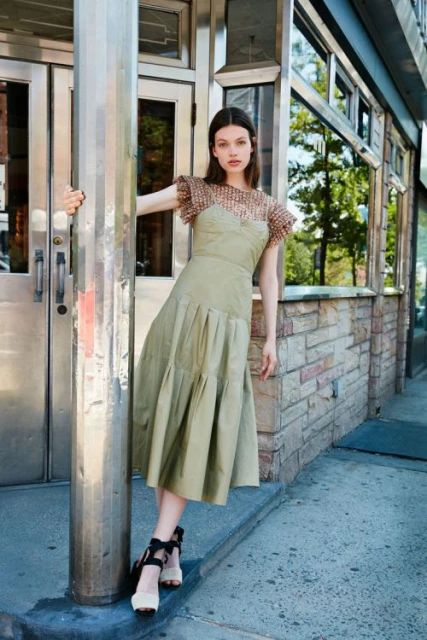 A young woman in a green dress poses in front of a building with metal doorframes. She is standing next to a silver pole.
