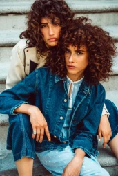 Two women with curly brown hair wearing denim clothing seated closely on stone steps with makeup by Mika Curella.