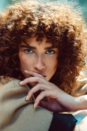 Close-up portrait of a woman with curly brown hair holding her hand near her face with metallic gold nail polish.