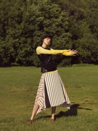 A woman in a black vest and striped skirt poses on a green lawn with trees in the background.