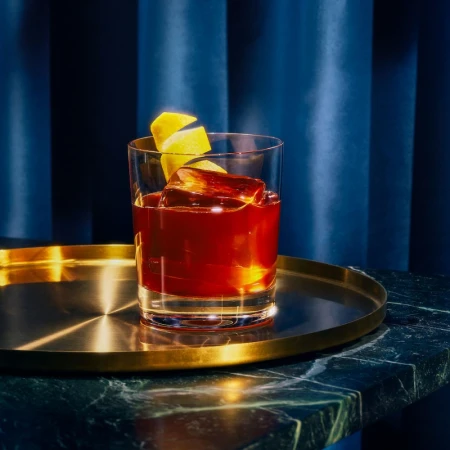A cocktail in a clear glass with a large ice cube and lemon peel, resting on a gold tray and dark green marble surface.